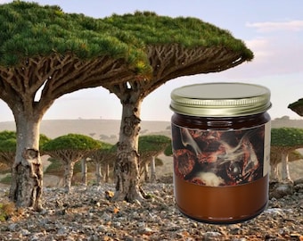 Può includere: Un barattolo di vetro con coperchio dorato, riempito con una sostanza marrone scuro, forse una candela o un balsamo. L'etichetta presenta un'immagine di legno che brucia. Lo sfondo mostra un paesaggio con alberi unici a forma di ombrello.