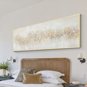Könnte beinhalten: Ein Schlafzimmer mit einem weißen Bett mit einer beigefarbenen Tagesdecke und einem geflochtenen Kopfteil. Das Bett ist mit weißen Bettwäsche bezogen und hat ein paar Kissen. Über dem Bett hängt ein großes abstraktes Gemälde mit goldener und weißer Farbe. Das Gemälde ist in Gold gerahmt. Neben dem Bett steht ein kleiner schwarzer Nachttisch.