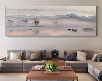 Puede incluir: Un sofá seccional gris con almohadas se encuentra frente a una gran pintura enmarcada de un paisaje montañoso brumoso con un reflejo de lago. La pintura está en tonos de gris, azul y rosa. Una mesa de café de madera con libros y una planta en maceta se encuentra frente al sofá.
