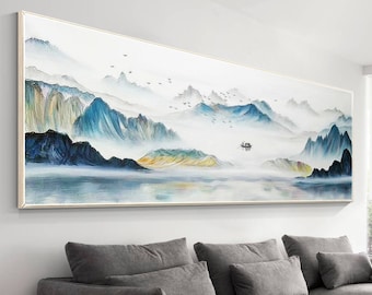 May include: A long, horizontal canvas painting depicting a misty mountain landscape with a small boat on a lake. The painting is framed in a thin, light wood frame and hangs on a white wall above a gray sectional sofa.