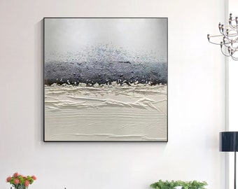 May include: A white and gray abstract painting with a textured surface. The painting is framed in a black frame and is hanging on a white wall. The painting is in a modern living room with a white table, black chairs, and a gray rug.