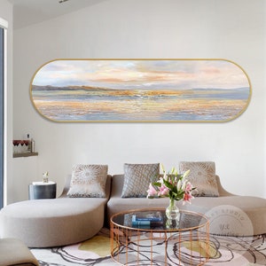 Puede incluir: Una pintura larga y horizontal sobre lienzo de un paisaje marítimo con una puesta de sol dorada. La pintura está enmarcada en un marco ovalado dorado y cuelga de una pared blanca sobre un sofá beige. La pintura es de estilo abstracto contemporáneo con una superficie texturizada.