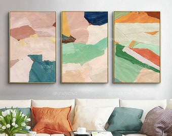 Op de afbeelding: Drie abstracte schilderijen in een moderne, minimalistische stijl. De schilderijen tonen een mix van roze, groen, blauw, oranje en witte kleuren. De schilderijen zijn ingelijst in goud.