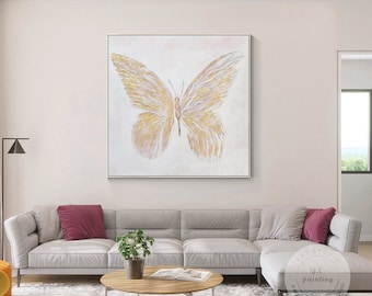 May include: A white canvas painting featuring a gold and pink butterfly in the centre. The painting is hanging on a white wall above a grey sofa.
