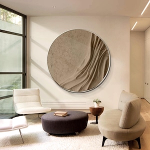 以下が含まれることがあります： テクスチャのあるベージュの表面と微妙な波模様が施された、大きな丸い壁掛けアート作品。アートはシルバーの金属フレームで囲まれ、モダンなリビングルームの白い壁に掛けられています。