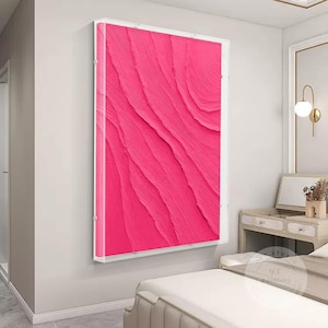 Puede incluir: Una pintura abstracta enmarcada con un diseño texturizado rosa. La pintura está en un marco blanco y está colgada en una pared blanca.
