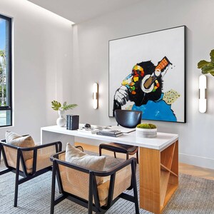 May include: A white desk with a wooden base and two black chairs with woven seats. A painting of a monkey wearing headphones hangs on the wall above the desk. The monkey is painted in a colorful style with blue, yellow, red, and green.