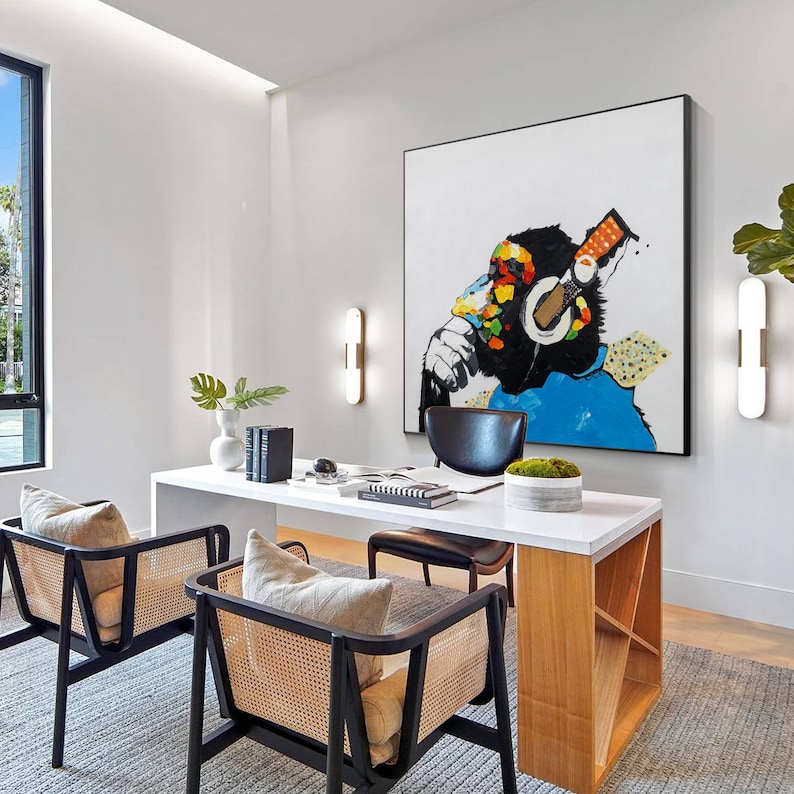 May include: A white desk with a wooden base and two black chairs with woven seats. A painting of a monkey wearing headphones hangs on the wall above the desk. The monkey is painted in a colorful style with blue, yellow, red, and green.