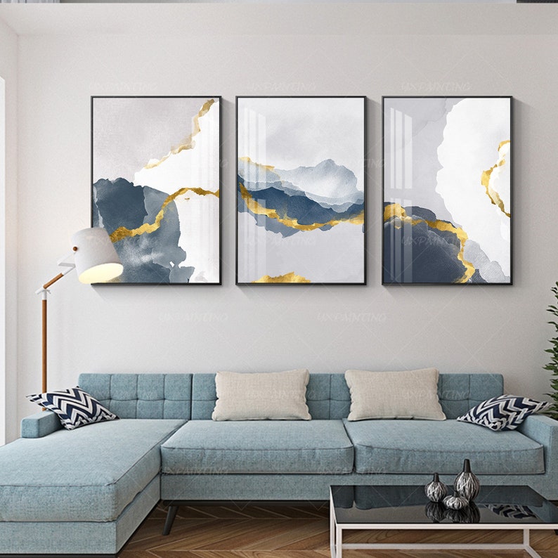 May include: Three abstract art prints with a grey and white background and gold accents. The prints are framed in black and are arranged in a row on a white wall.