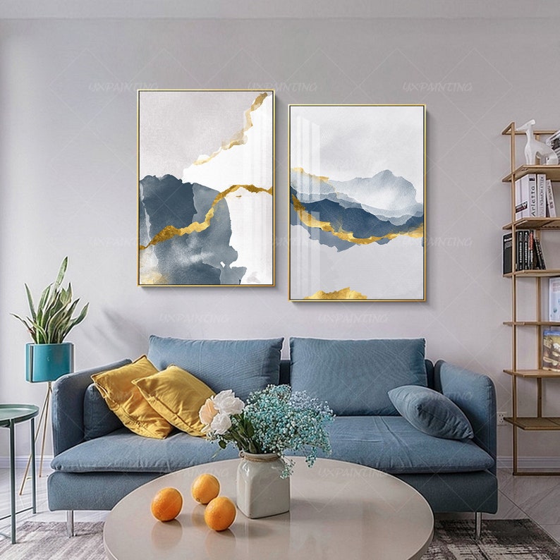 May include: Two abstract paintings with gold accents on a white background. The paintings are framed in gold and are hanging on a white wall above a blue sofa.