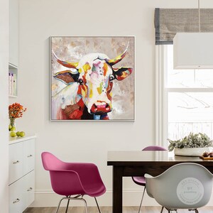 May include: A colorful abstract painting of a cow's head with a white background. The cow's fur is painted in shades of red, yellow, blue, and green. The painting is framed in a white frame.