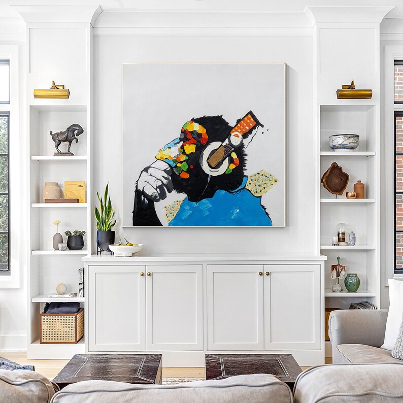May include: A colorful abstract painting of a monkey wearing headphones. The monkey is sitting with its chin on its hand and looking to the side. The painting is on a white wall above a white cabinet with three doors.
