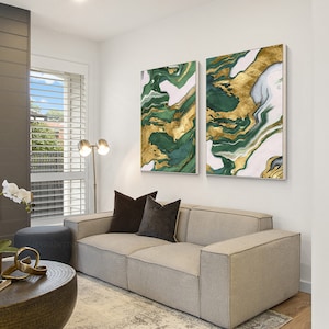 May include: Two rectangular wall art canvases featuring an abstract marbled pattern in shades of deep forest green, white, and metallic gold. The paintings are displayed side-by-side on a plain white wall above a light gray upholstered sofa.