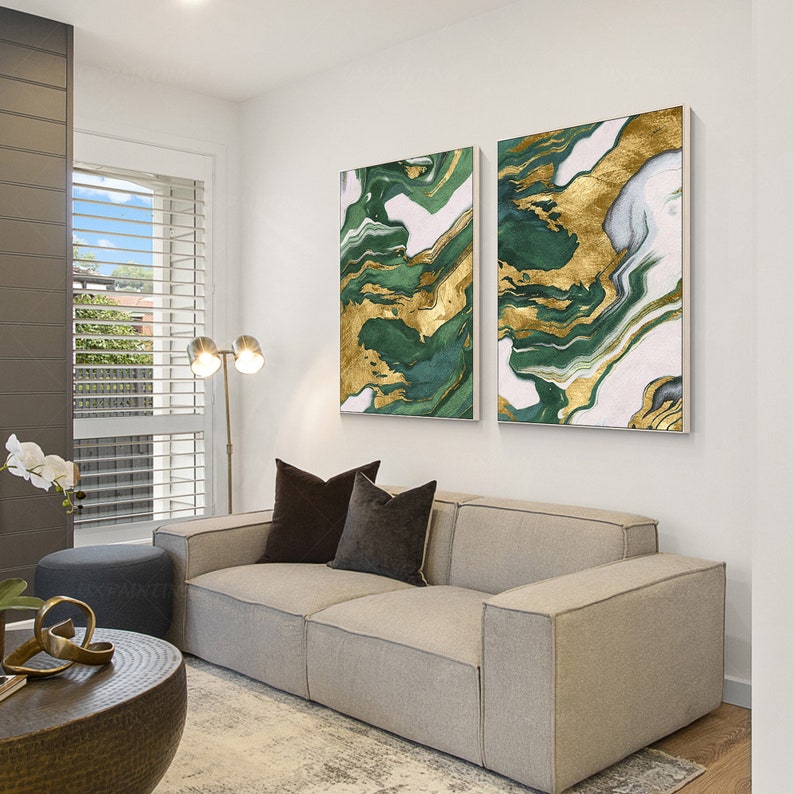 May include: Two rectangular wall art canvases featuring an abstract marbled pattern in shades of deep forest green, white, and metallic gold. The paintings are displayed side-by-side on a plain white wall above a light gray upholstered sofa.