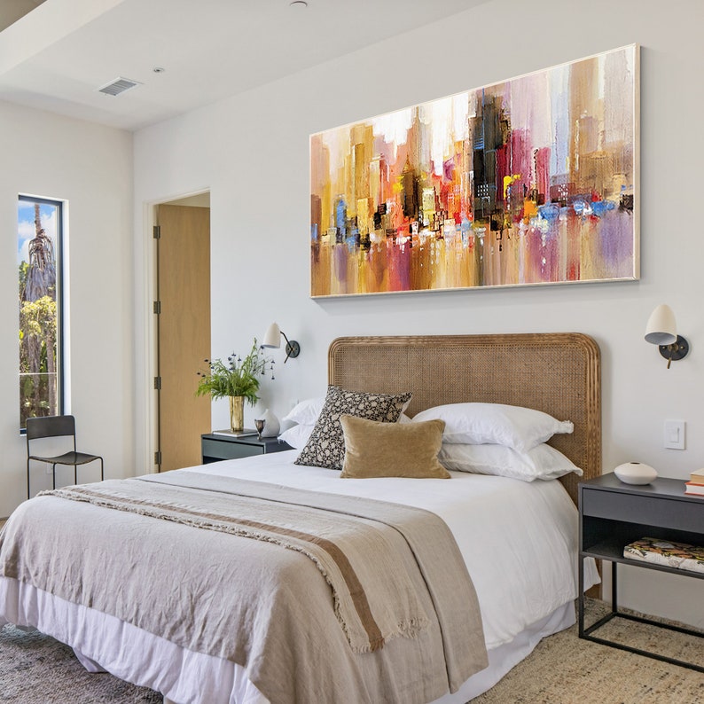 May include: A bedroom with a large abstract painting of a city skyline above a bed with a beige headboard and a white duvet cover. The bed has a beige throw blanket and a few pillows. There is a small black side table next to the bed.