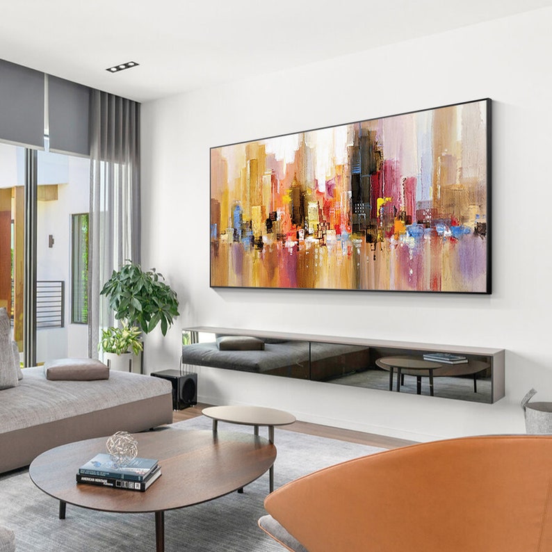 May include: Abstract cityscape painting with a mix of yellow, orange, red, blue, and purple hues. The painting is framed in black and hangs on a white wall above a black entertainment center.