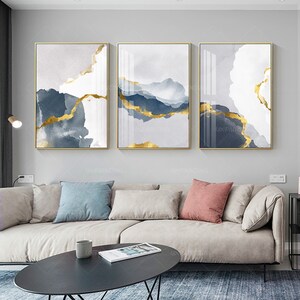 May include: Three abstract art prints with gold accents on a white background. The prints are framed in gold and are hanging on a grey wall above a white sofa.