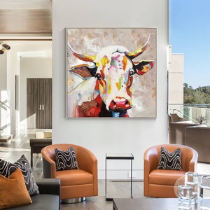 May include: A colorful abstract painting of a cow's head with a white background. The cow's face is painted in bright colors, including red, yellow, blue, and green. The painting is framed in a white frame.