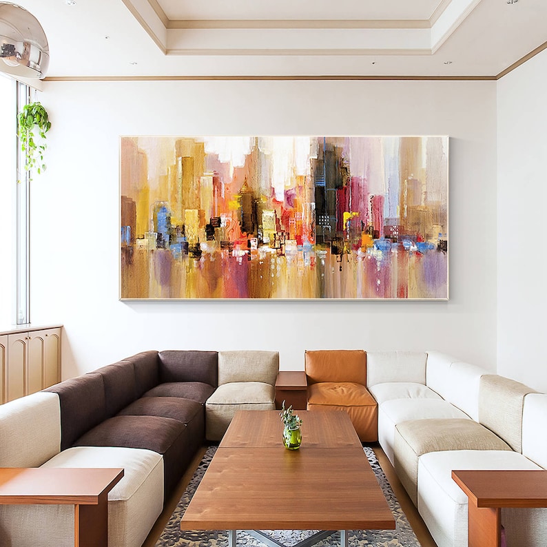 May include: Abstract painting of a city skyline in shades of brown, yellow, red, blue, and purple. The painting is framed and hanging on a white wall above a brown and beige sectional sofa.