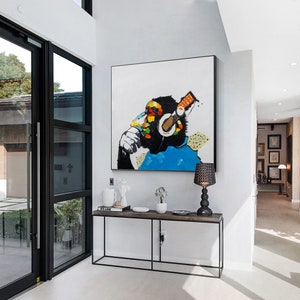 May include: A colorful abstract painting of a monkey wearing headphones and holding a microphone. The monkey is painted in shades of black, blue, yellow, red, and green. The painting is framed and hanging on a white wall.