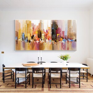 May include: Abstract cityscape painting in warm colors, featuring a mix of yellow, orange, red, and blue hues. The painting is displayed above a long, white dining table with black chairs.