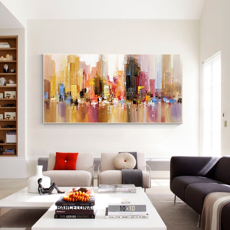 May include: Abstract painting of a cityscape with colorful buildings and a reflection of the city in the water. The painting is in a modern living room setting with a white coffee table and two beige sofas.