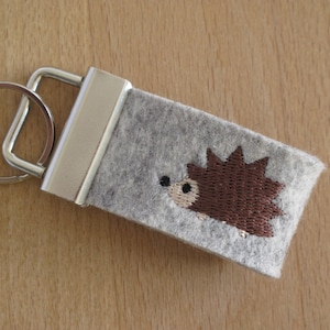 Könnte beinhalten: Ein grauer Filz-Schlüsselanhänger mit einem braunen, gestickten Igel-Design. Der Schlüsselanhänger hat einen silbernen Metallverschluss und Schlüsselring. Der Igel hat ein beiges Gesicht und schwarze Augen. Der Hintergrund ist eine hellbraune Holzoberfläche.