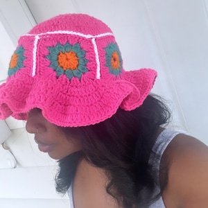 May include: A pink crocheted bucket hat with white trim and colorful flower designs. The hat has a wide brim and a ruffled edge.