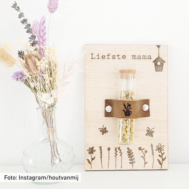 Peut inclure: Une plaque en bois avec le texte "Liefste mama" et un petit tube en verre rempli de graines. La plaque comporte &eacute;galement une petite maison d'oiseau et une sangle en cuir avec une boucle en m&eacute;tal. La plaque est entour&eacute;e d'un fond blanc.