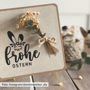 May include: A handmade Easter card with a beige background and a brown border. The card features a black bunny illustration with the words "Frohe OSTERN". A small bouquet of dried flowers and greenery is tied to the card with twine.