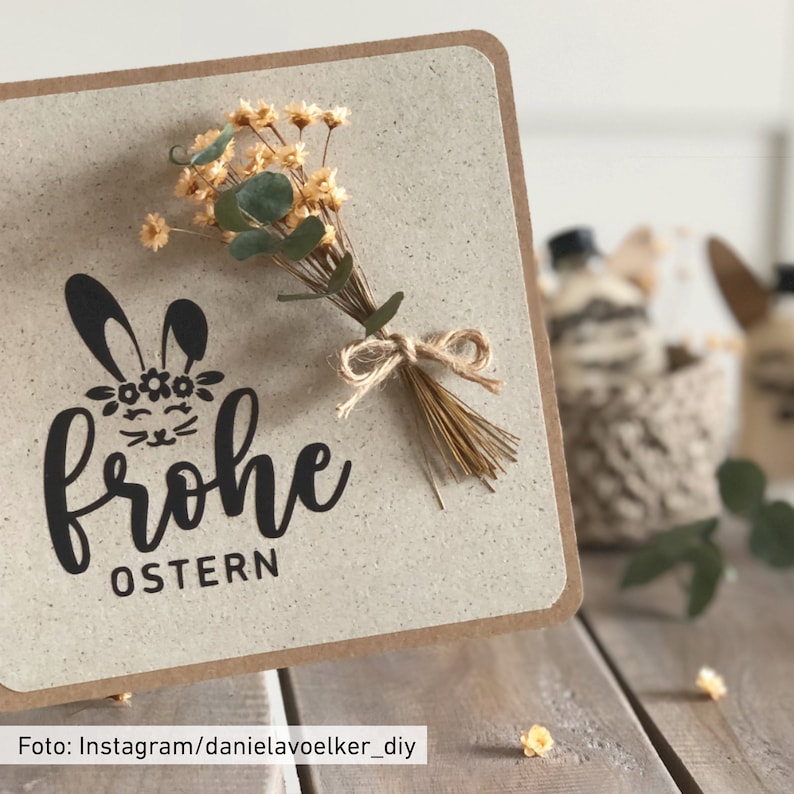 May include: A handmade Easter card with a beige background and a brown border. The card features a black bunny illustration with the words "Frohe OSTERN". A small bouquet of dried flowers and greenery is tied to the card with twine.
