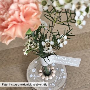 Peut inclure: Un petit bouquet de fleurs blanches et roses dans un vase en verre avec une base en bois. Le vase est d&eacute;cor&eacute; d'une cha&icirc;ne de perles en bois et la base a un motif floral. Une &eacute;tiquette avec le texte "HELLO Spring" est attach&eacute;e au vase.