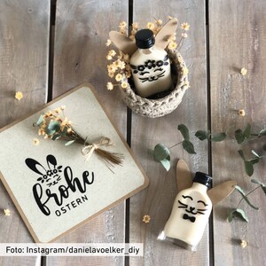 May include: Easter-themed decorations. A bottle decorated with a bunny face and ears, a greeting card with the words "Frohe Ostern", and a small woven basket with a bottle inside. The items are on a wooden surface.