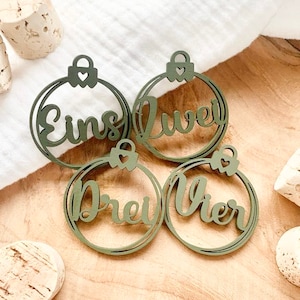 May include: Four green wooden Christmas ornaments with the German words "Eins", "Zwei", "Drei", and "Vier" written on them.