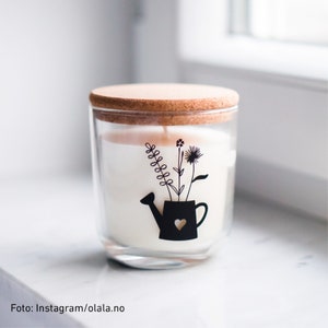 Peut inclure: Un bocal en verre pour bougie avec un couvercle en li&egrave;ge et un motif noir et blanc d'un arrosoir avec des fleurs. La bougie est blanche et le bocal est transparent.