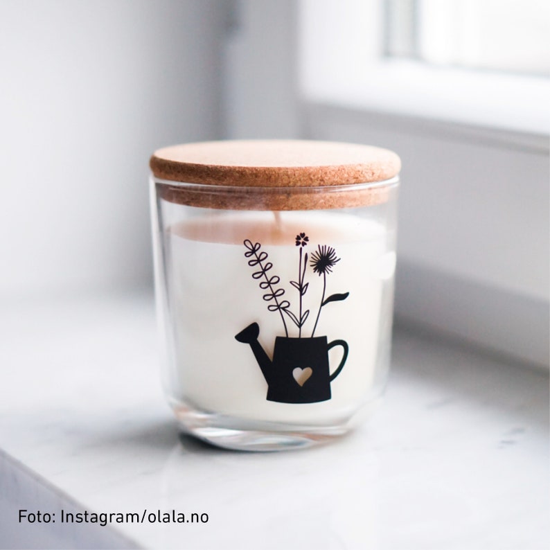 Peut inclure: Un bocal en verre pour bougie avec un couvercle en li&egrave;ge et un motif noir et blanc d'un arrosoir avec des fleurs. La bougie est blanche et le bocal est transparent.