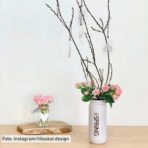 Puede incluir: Un jarr&oacute;n blanco con el texto "HEJ SPRING" en &eacute;l, lleno de rosas rosas y ramas con huevos decorativos colgantes. Tambi&eacute;n se puede ver un tarro de vidrio m&aacute;s peque&ntilde;o con un dise&ntilde;o de coraz&oacute;n, lleno de rosas rosas.