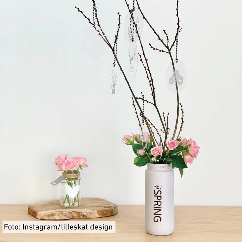 Puede incluir: Un jarr&oacute;n blanco con el texto "HEJ SPRING" en &eacute;l, lleno de rosas rosas y ramas con huevos decorativos colgantes. Tambi&eacute;n se puede ver un tarro de vidrio m&aacute;s peque&ntilde;o con un dise&ntilde;o de coraz&oacute;n, lleno de rosas rosas.