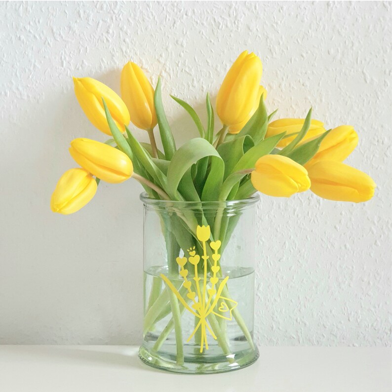 Peut inclure: Un vase en verre transparent avec un motif floral jaune pos&eacute; sur une surface blanche. Le vase est rempli de tulipes jaunes.
