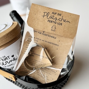 May include: A gift basket containing a heart-shaped cookie cutter, a package with German text, and a jar with text. The package reads "AUF DIE Plätzchen FERTIG LOS" and includes a recipe. The basket is black with a wooden handle.