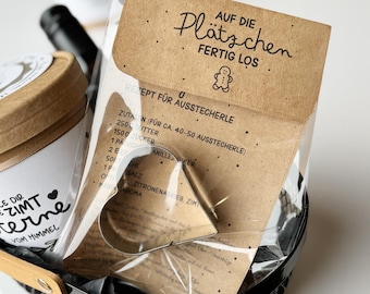 May include: A gift basket containing a heart-shaped cookie cutter, a package with German text, and a jar with text. The package reads "AUF DIE Plätzchen FERTIG LOS" and includes a recipe. The basket is black with a wooden handle.