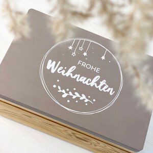 May include: A gray wooden box with a hinged lid. The lid has a white circle with the text "Frohe Weihnachten" and a small branch with leaves.