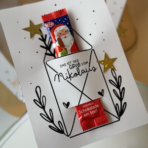 May include: A white card with a black line drawing of an envelope, containing a Santa Claus chocolate bar and a red chocolate on a stick. The card has gold star stickers and the text "DAS IST DER GRUß VOM Nikolaus".