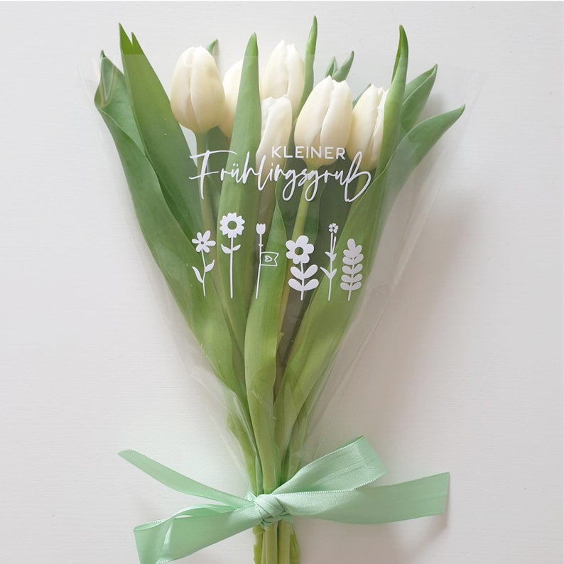 Peut inclure: Un bouquet de tulipes blanches envelopp&eacute; dans du cellophane transparent avec un ruban vert. Le bouquet porte une &eacute;tiquette blanche avec le texte "KLEINER Fr&uuml;hlingsgru&szlig;" et un petit graphique de fleurs.