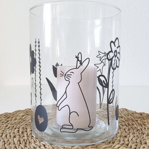 Puede incluir: Un portavelas de vidrio transparente con un dise&ntilde;o en blanco y negro de un conejo, flores y corazones. El portavelas est&aacute; sobre una superficie tejida.