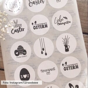 Puede incluir: Una hoja de pegatinas redondas blancas con dise&ntilde;os de Pascua negros. Los dise&ntilde;os incluyen conejos, huevos, flores y texto en alem&aacute;n e ingl&eacute;s. El texto incluye "Feliz Pascua", "Frohe Ostern", "Liebe Ostergr&uuml;&szlig;e", "Hasenpost" y "Kunterbunte Ostern".