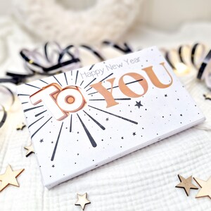 Pode incluir: Um cartão branco com as palavras "To You" em um design dourado e branco, com as palavras "Happy New Year" acima. O cartão tem um design de explosão de estrelas e pequenos detalhes de estrelas. Fitas pretas e douradas e estrelas de madeira no fundo.