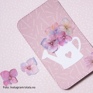 Peut inclure: Une carte rose avec un arrosoir blanc et des fleurs roses. La carte a une bordure blanche cousue et un fond &agrave; pois roses.