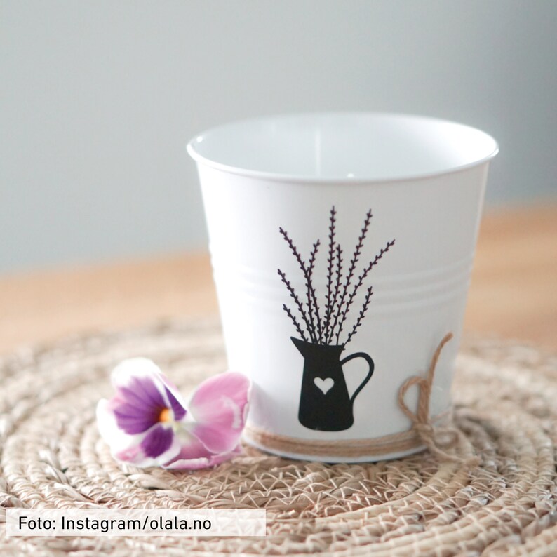 Puede incluir: Cubo de metal blanco con una silueta negra de un jarr&oacute;n con flores de lavanda. El cubo est&aacute; sentado en un mantel individual tejido con una flor morada y rosa en primer plano.
