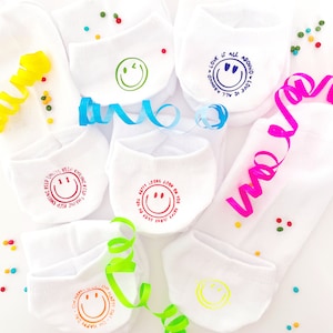 Könnte beinhalten: Sechs weiße Socken mit verschiedenen bunten Smiley-Gesichtern und Sprüchen darauf gedruckt. Die Sprüche lauten: "Liebe ist überall", "Glücklich aussehen steht dir gut", "Lächeln hilft" und "Oh, glücklicher Tag".